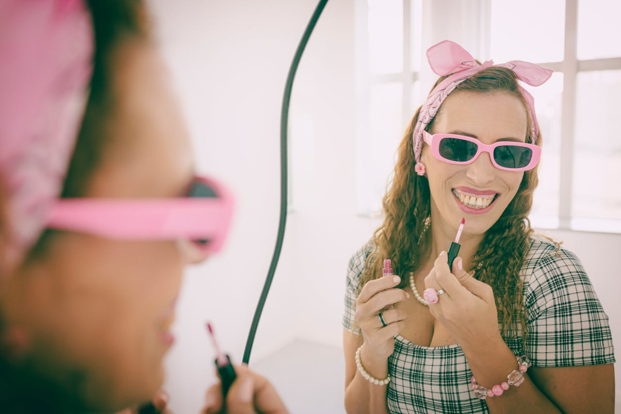 Cheerful woman in pink accessories applies lipstick in front of a mirror, exuding a vintage vibe.