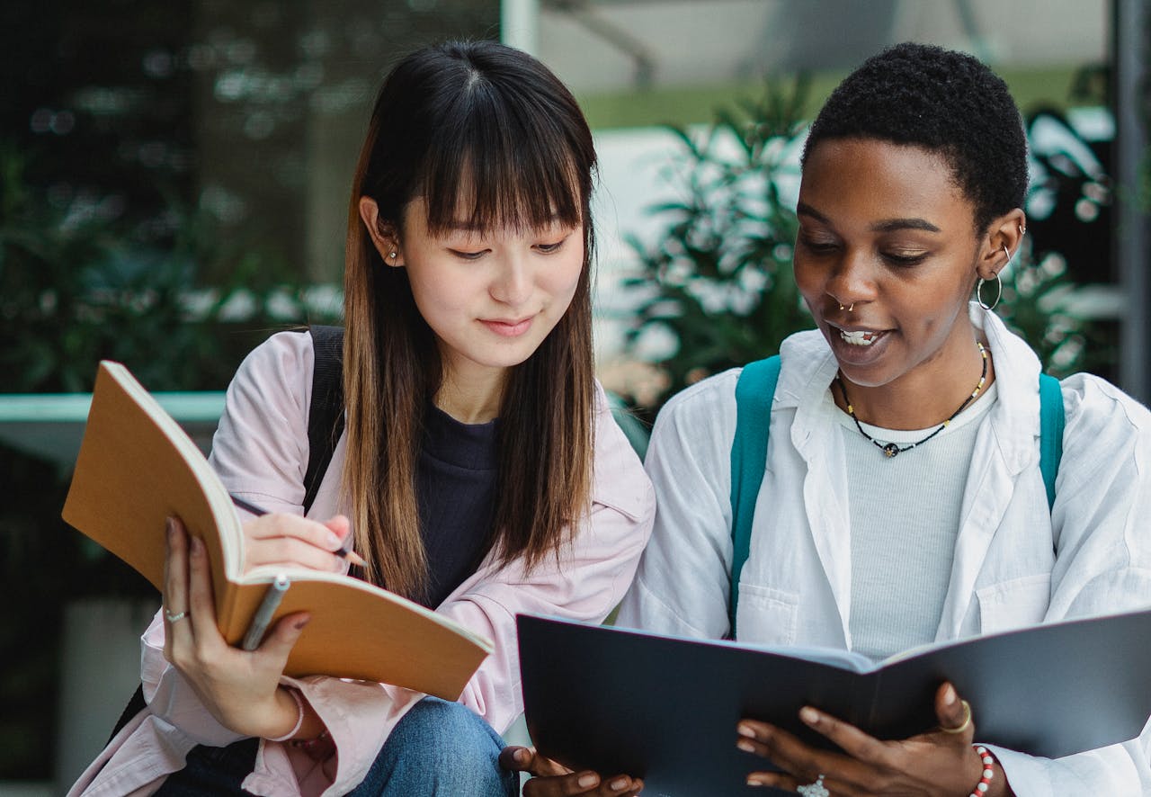 our-story-01 Young smiling black female student with exercise book talking to Asian partner on street in daylight