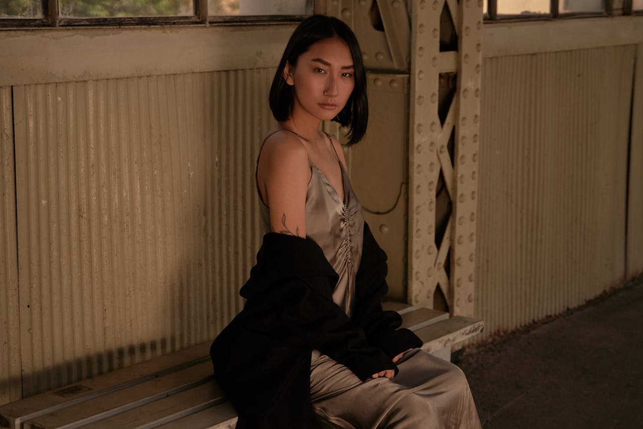 An elegant woman in a silk dress poses stylishly on a wooden bench indoors.